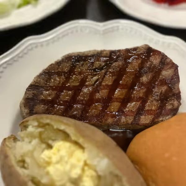 Charbroiled steak with grill marks, a baked potato with butter, and a dinner roll on a white plate.